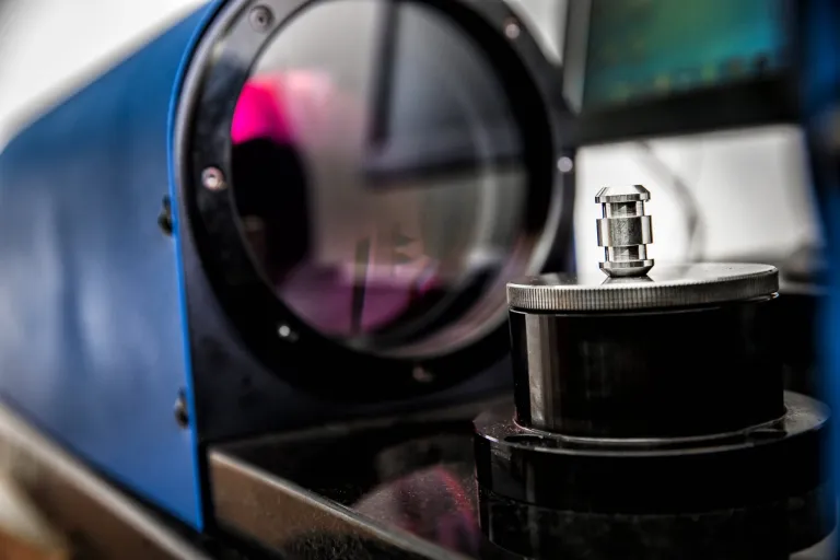 A small machined part in front of a blue measuring machine lens