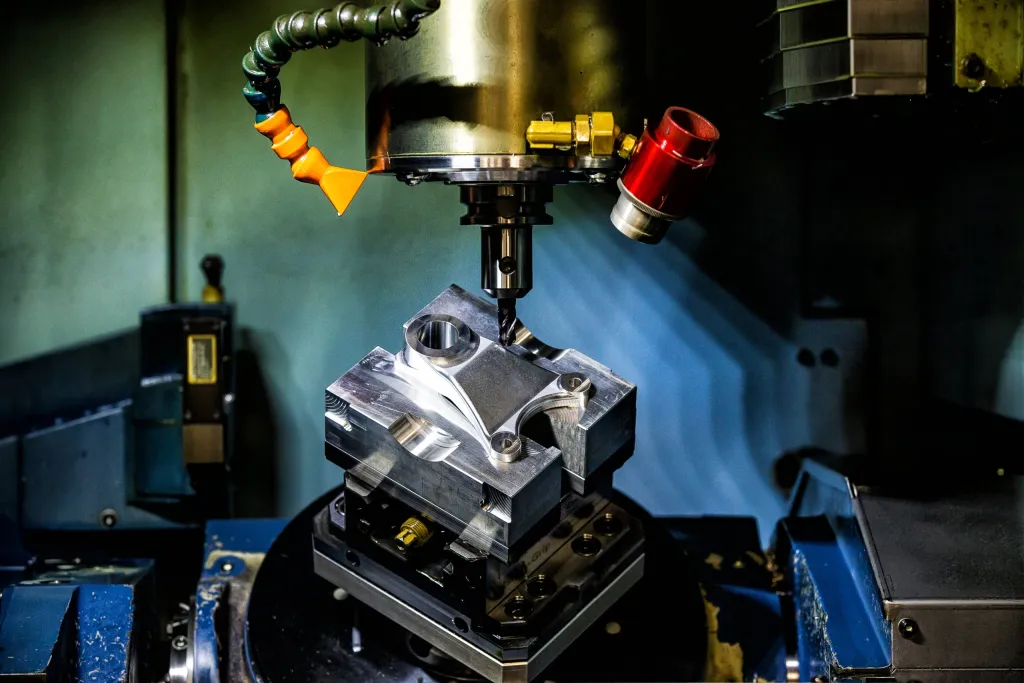 A machined metal part held in a fixture beneath a CNC milling spindle, coolant nozzles in view